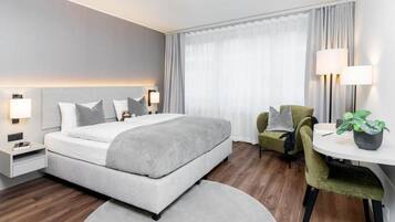 Camera d'hotel moderna con un letto grande, lampade ai comodini, una poltrona verde, un piccolo tavolino con una pianta e vista all'esterno tramite tende trasparenti.