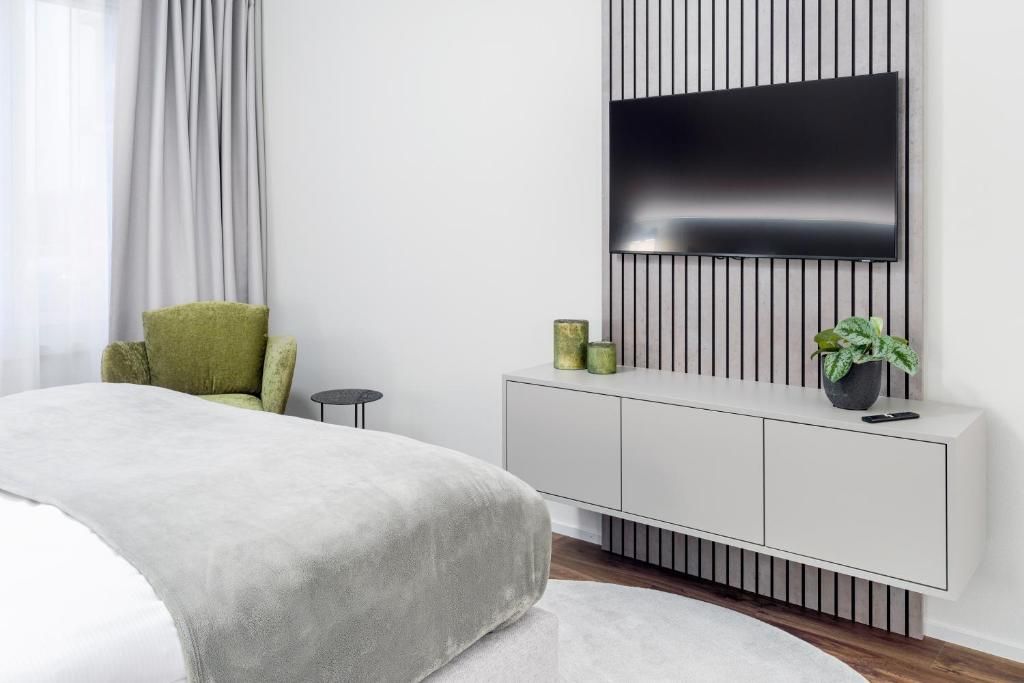 A modern bedroom with a bed, a TV mounted on a wall, a green chair, a small table, and a cabinet with decorative items.
