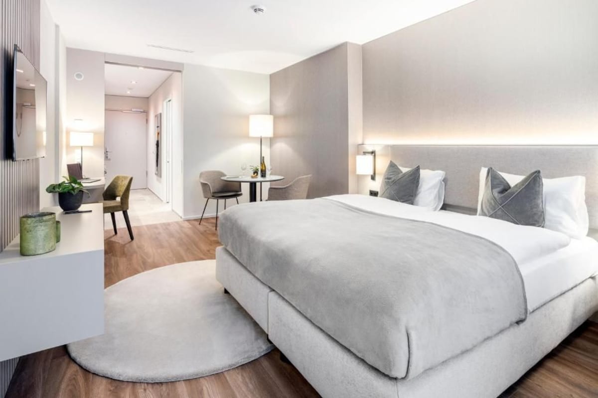 A modern hotel room with a large bed, a desk, a chair, and a small table with a lamp.