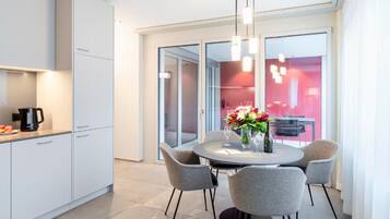 A modern kitchen with white cabinetry, a dining area with a round table and grey chairs, and a large window with a red interior view.