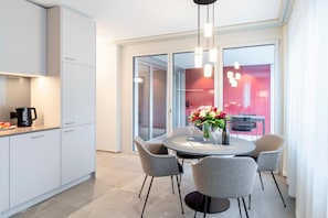 Una cocina moderna con armarios blancos, un comedor con una mesa redonda y sillas grises, y un amplio ventanal con vista al interior en tonos rojos.