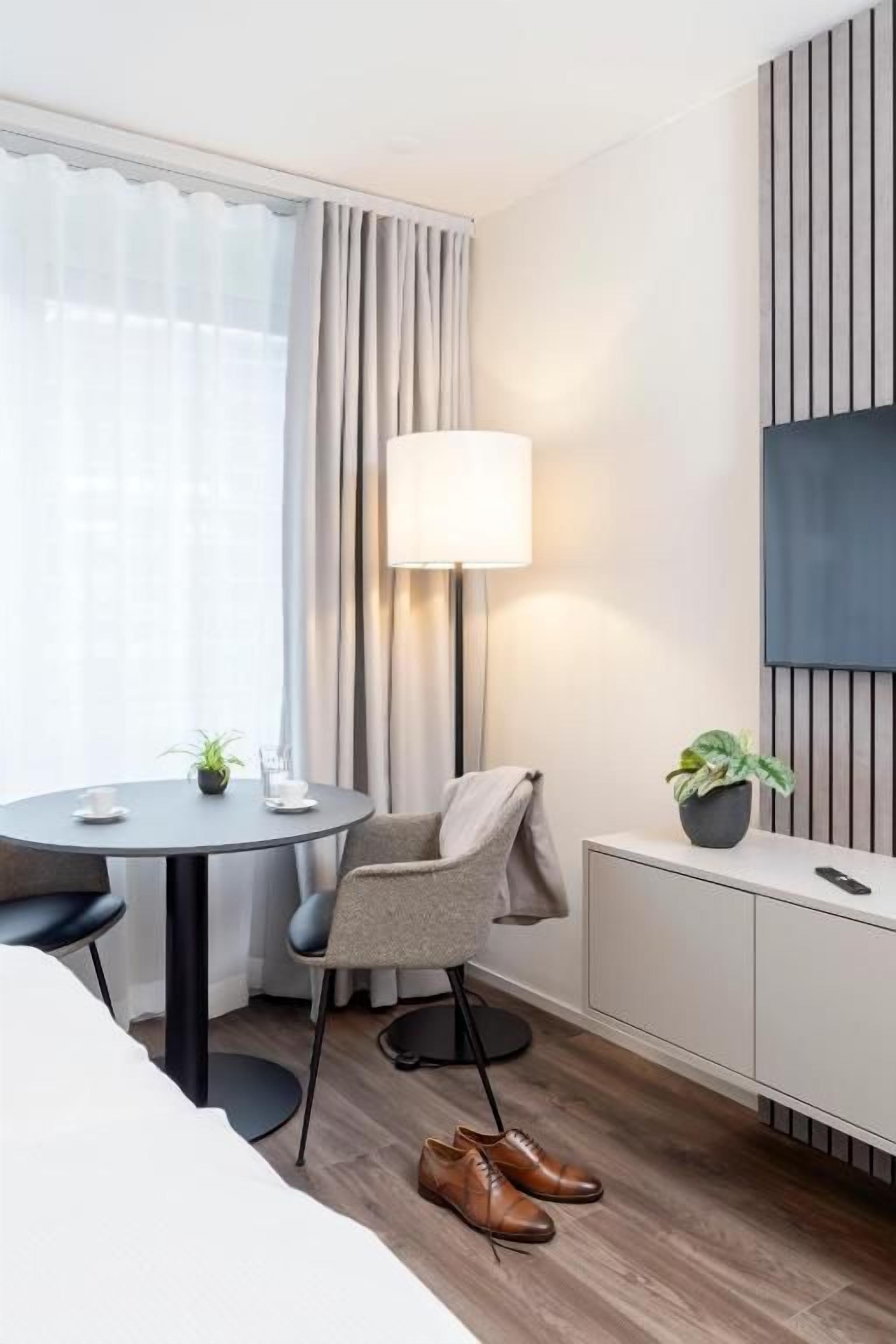 Habitación de hotel con una cama, un escritorio con lámpara, una silla, una planta en maceta y un televisor sobre un pedestal.