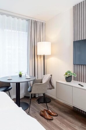 A hotel room with a bed, a desk with a lamp, a chair, a potted plant, and a TV on a stand. - CITY STAY - Papieri-Ring (Cham)
