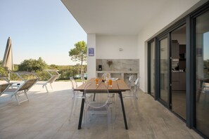 A modern outdoor dining area with a wooden table and clear plastic chairs, a poolside lounge with sun loungers, and a kitchen visible through glass doors. - New Listing (Krnica)