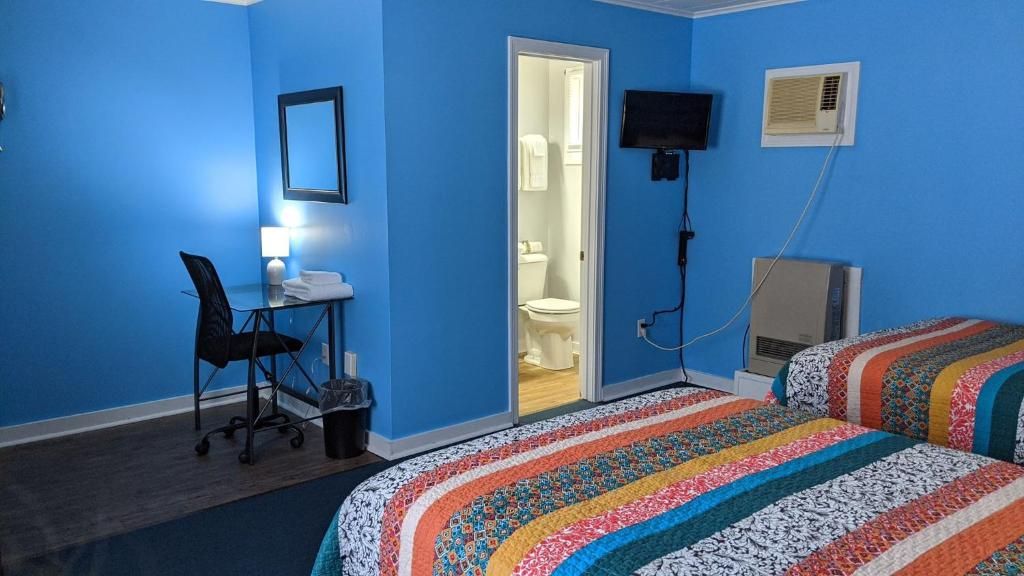 A room with a blue wall, a desk with a chair, a bed with a colorful blanket, a television, and an air conditioning unit.