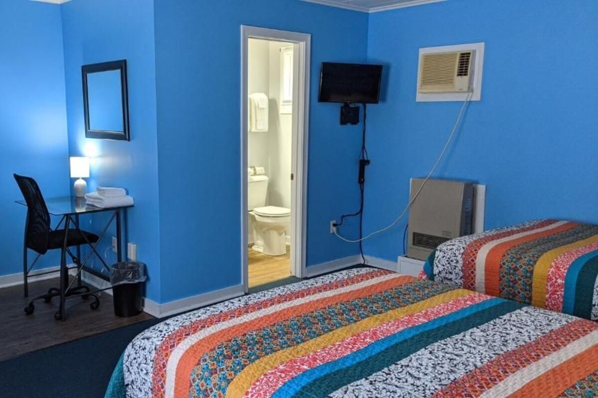 A room with a blue wall, a desk with a chair, a bed with a colorful blanket, a television, and an air conditioning unit.