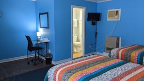 A room with a blue wall, a desk with a chair, a bed with a colorful blanket, a television, and an air conditioning unit. - Blue Ridge Motel (Schroon Lake)