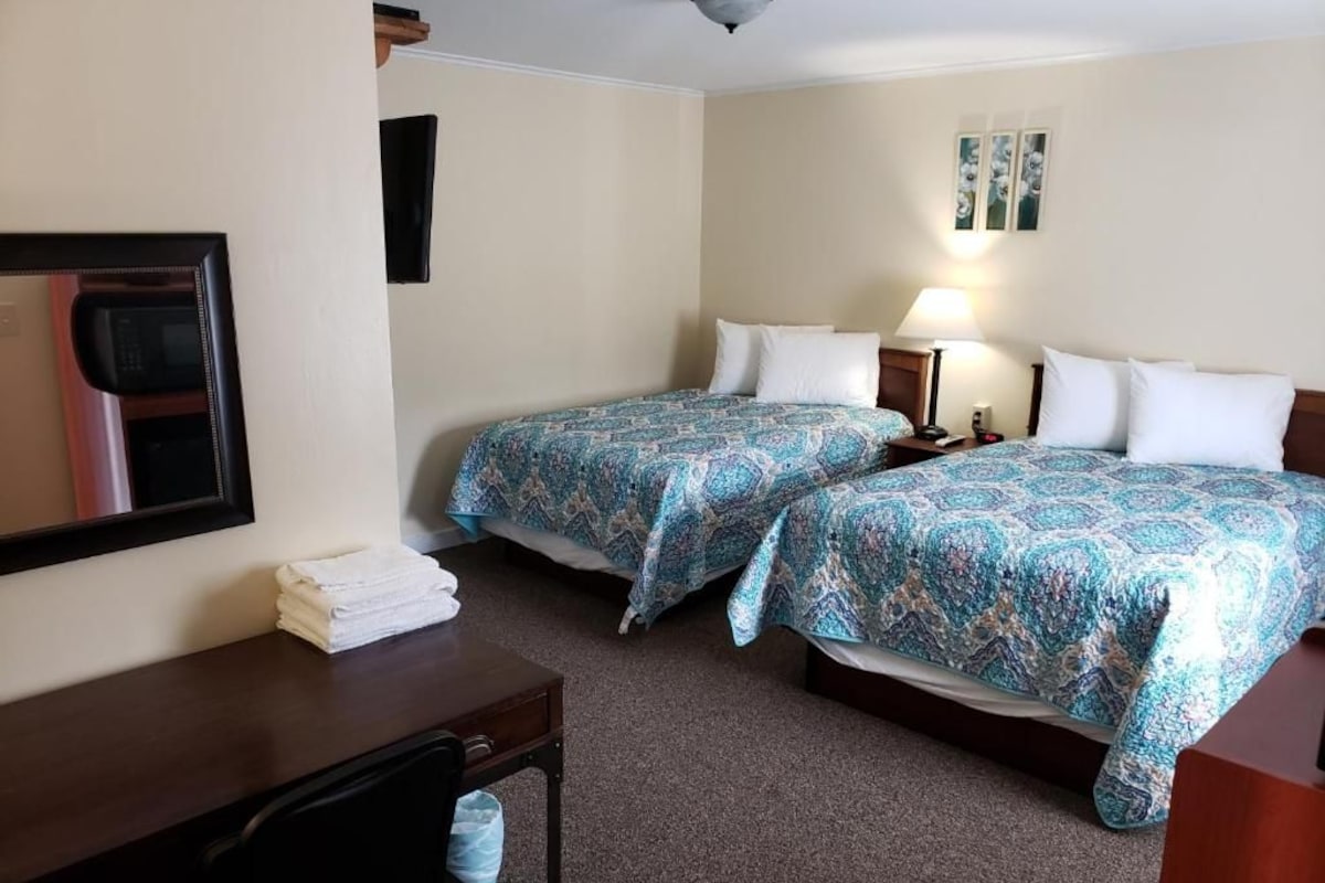 A hotel room with two beds, a desk, a chair, a microwave, and a wall-mounted TV.