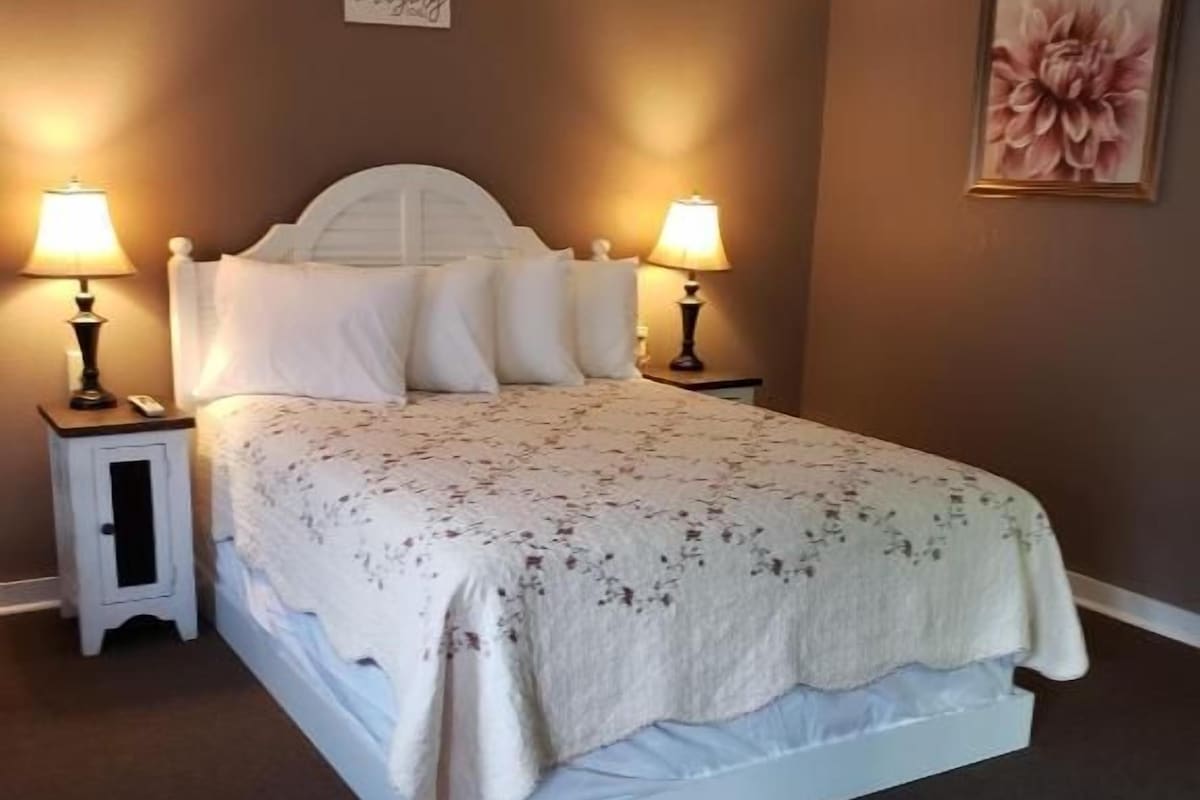 A bedroom with a bed, two bedside tables, a nightstand, a wall-mounted sign, and a framed picture.