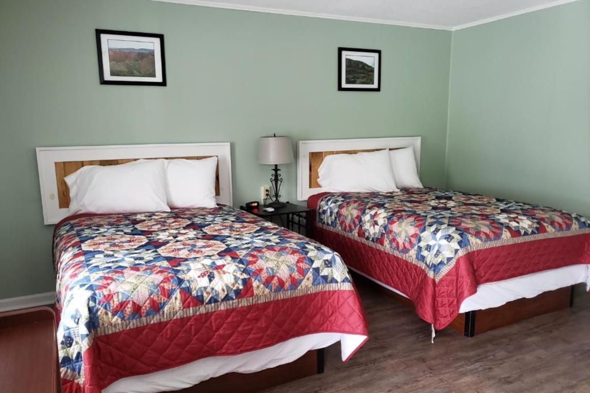 A room with two beds, each with a quilted bedspread and white pillows. There are two framed pictures on the wall, and a bedside table with a lamp.