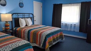 A bedroom with two beds, a nightstand with a lamp, a door, and a window with curtains. - Blue Ridge Motel (Schroon Lake)