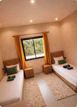 A hotel room with two beds, a window with curtains, a small bedside table with a lamp, and a view of greenery outside. - New Listing (Dar El Hamra)