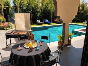 An outdoor dining area with a pool view, a grill, and a table set with drinks and plates. - New Listing (Dar El Hamra)