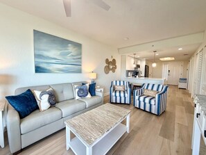 A living room with a sofa, armchairs, and a coffee table. There is a kitchen in the background. - Bright Beachfront Penthouse | Balcony Views + Pool (Treasure Island)
