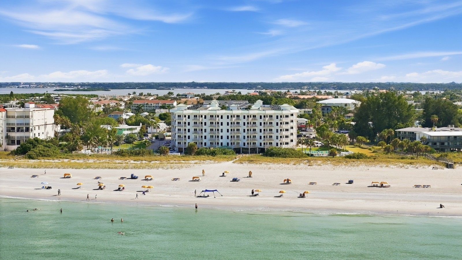 Un hotel frente a la playa con varios pisos, una playa arenosa en primer plano y vista al océano y al horizonte urbano.