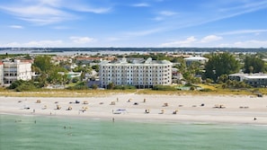 A beachfront hotel with multiple floors, a sandy beach in the foreground, and a view of the ocean and distant cityscape. - Beachfront Escape Sunset Vista Resort Views & Pool (Treasure Island)