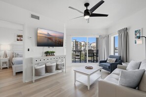 A modern living room with a flat-screen TV, a ceiling fan, a sofa, a coffee table, and a balcony with a view of buildings. - Beachfront Escape Sunset Vista Resort Views & Pool (Treasure Island)