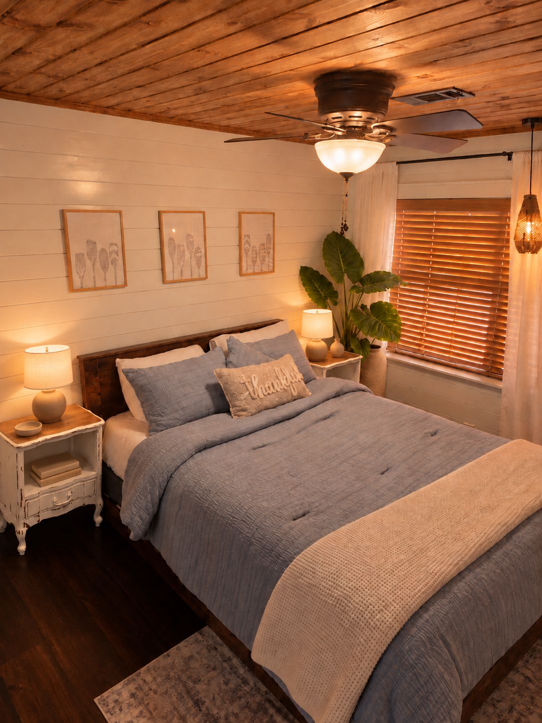 A bedroom with a wooden ceiling, a bed with blue bedding, a nightstand with a lamp, and a large plant.