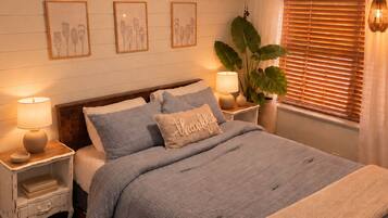 A bedroom with a wooden ceiling, a bed with blue bedding, a nightstand with a lamp, and a large plant.