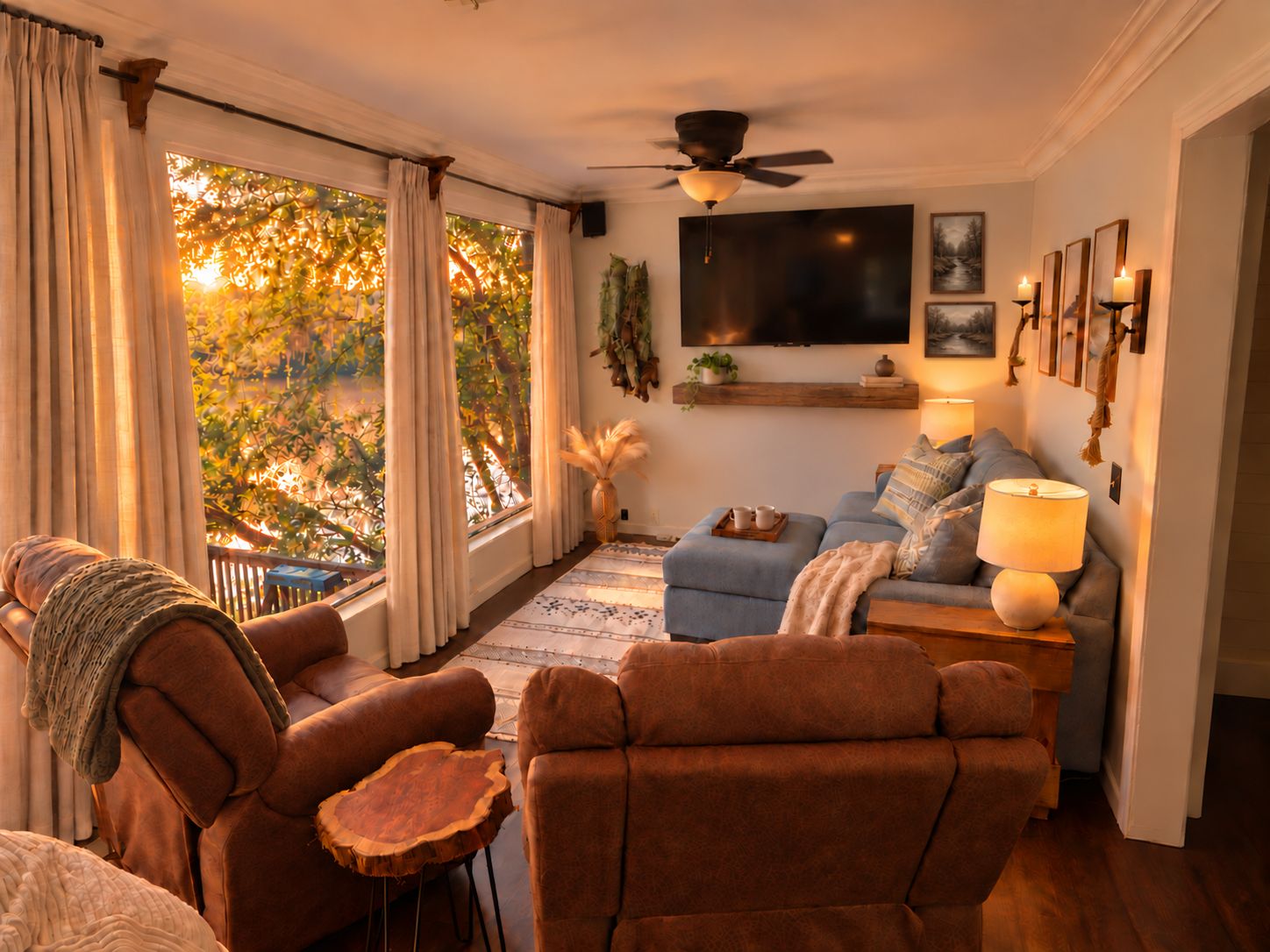 A cozy living room with a sofa, armchairs, and a coffee table. There is a television mounted on the wall, a ceiling fan, and a window with curtains.