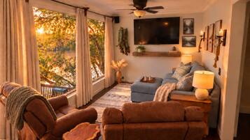 A cozy living room with a sofa, armchairs, and a coffee table. There is a television mounted on the wall, a ceiling fan, and a window with curtains.