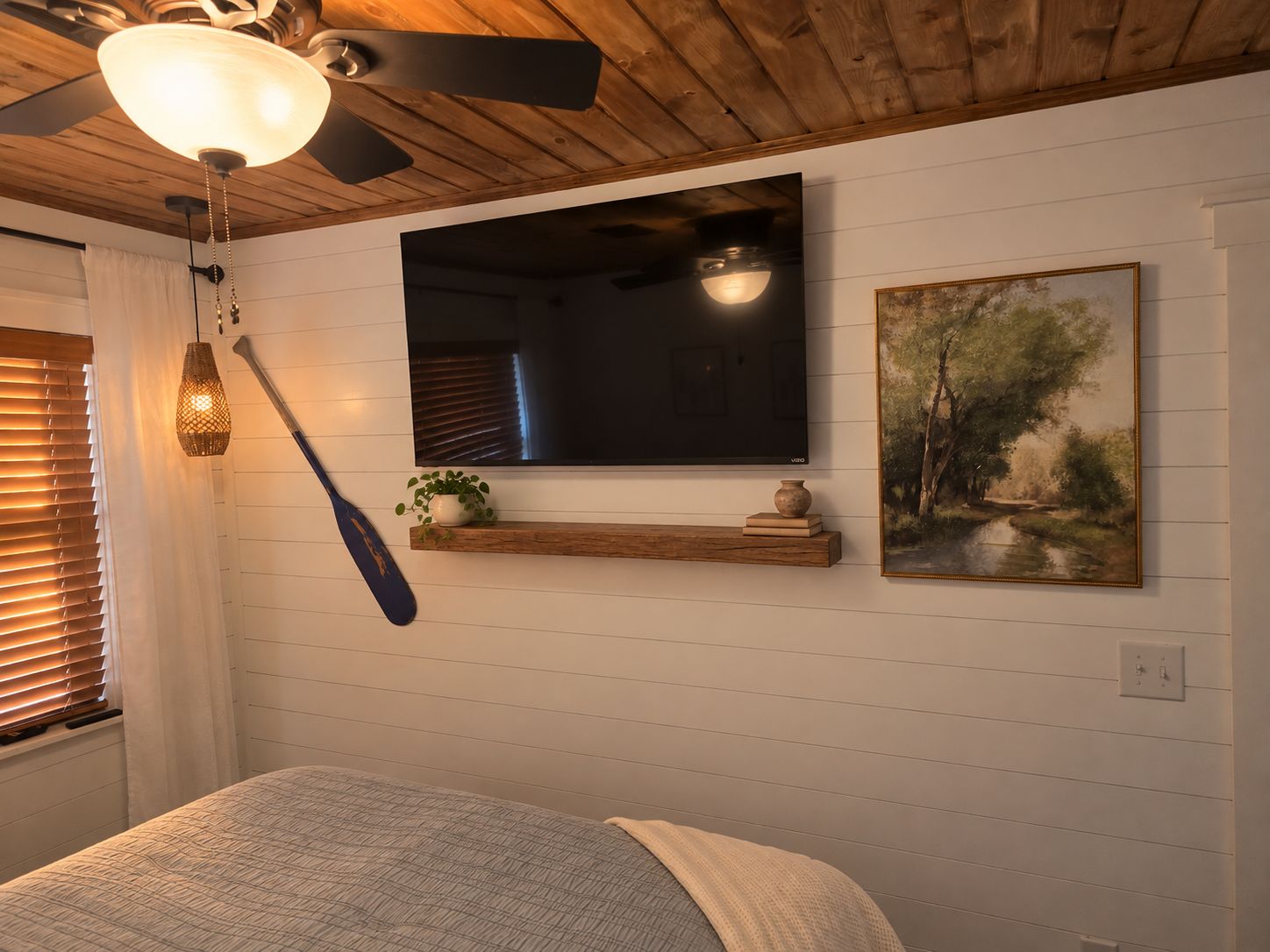Un dormitorio con cama, televisor montado, un cuadro, un estante de madera con adornos y un ventilador de techo.