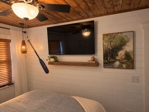A bedroom with a bed, a mounted TV, a painting, a wooden shelf with decorative items, and a ceiling fan.