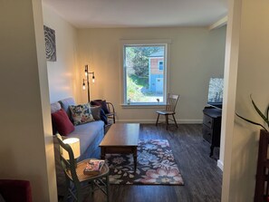 Un salon comprenant un canapé, une table basse, un fauteuil, un tapis, une lampe, un téléviseur et une fenêtre donnant sur une maison.