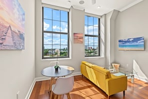 A modern living room with large windows, a yellow sofa, a dining table with chairs, and artwork on the walls. - Polished Metro 1BD Retreat | Top Eats | Sleeps 2 (Philadelphia)