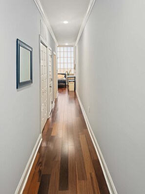 A hallway with a wooden floor, white walls, and a door leading to a room with a bed and a window. - Prime Studio City Hideaway | Gym Pass | Fits 2 (Philadelphia)