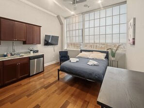 A modern bedroom with a large window, a bed with towels, a TV, and a kitchen area with wooden cabinets and a dishwasher. - Prime Studio City Hideaway | Gym Pass | Fits 2 (Philadelphia)
