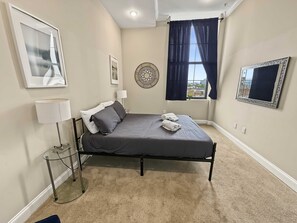 Ein ordentlich eingerichtetes Schlafzimmer mit einem Bett, einer Nachttischlampe, einem Wandventilator und einem Fenster mit Vorhängen.