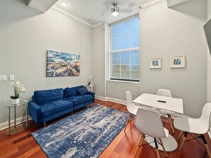 Ein modernes Wohnzimmer mit einem blauen Sofa, einem weißen Esstisch mit Stühlen, einem großen Fenster und einem Stadtbild an der Wand.