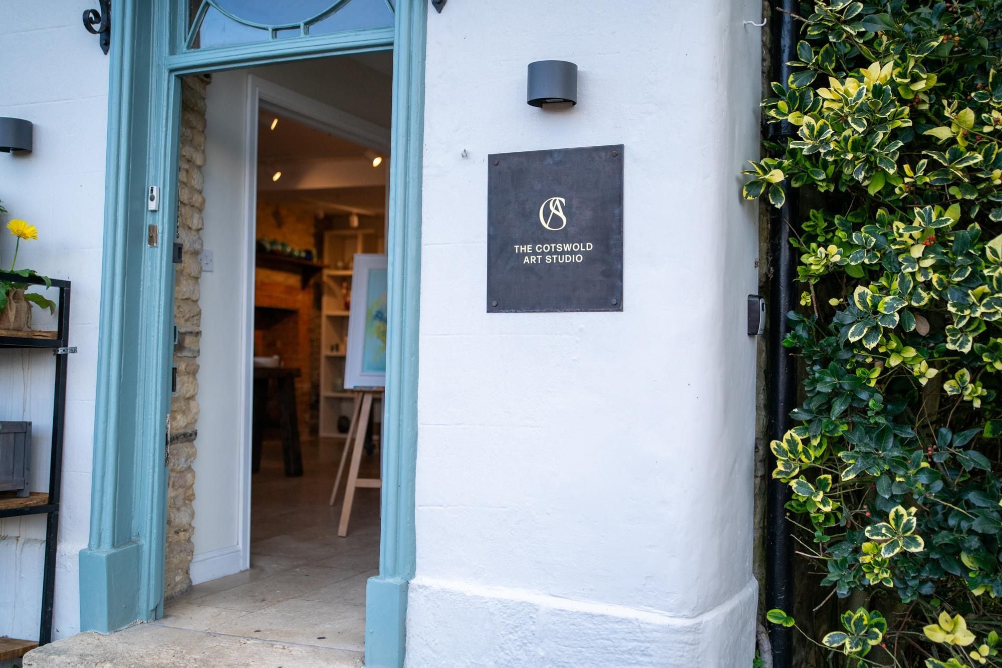 The entrance to The Cotswold Art Studio, featuring a stone wall, a metal door frame, and a signboard.
