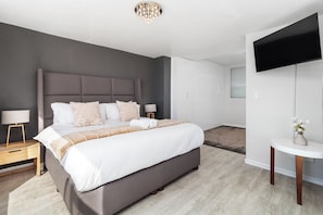 A modern bedroom with a large bed, a flat-screen TV mounted on the wall, and a small bedside table with a lamp. - Spacious Beachfront Retreat | Stunning Ocean Views (Cape Town)