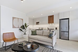 A modern living room with a sofa, coffee table, and a dining area with a kitchen in the background. - Luxe Modern Apartment | Ocean Views | Aircon (Umhlanga)