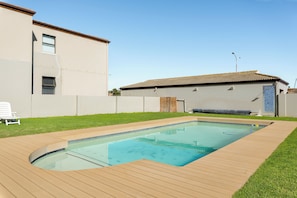 A rectangular swimming pool with a wooden deck surrounding it, set against a backdrop of a two-story building with a tiled roof. - Stylish 2 Bed Apartment | Big Bay Beach Club (Cape Town)