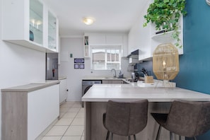A modern kitchen with white cabinetry, a central island with bar stools, a built-in dishwasher, and a hanging plant. - Stylish 2 Bed Apartment | Big Bay Beach Club (Cape Town)