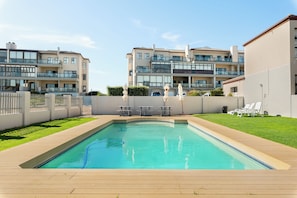 A rectangular swimming pool with a wooden deck, surrounded by a low fence and a grassy area. There are lounge chairs and tables in the background. - Stylish 2 Bed Apartment | Big Bay Beach Club (Cape Town)
