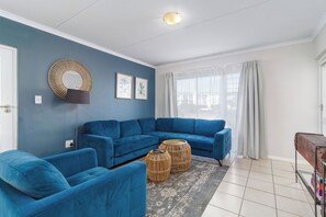 A living room with a blue sofa set, a round mirror, two framed botanical prints, a wicker side table, and a window with sheer curtains. - Stylish 2 Bed Apartment | Big Bay Beach Club (Cape Town)