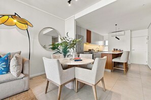 Un salon moderne avec une table à manger ronde, des chaises recouvertes d’un tissu gris, un canapé agrémenté de coussins à motifs, un grand miroir et un luminaire suspendu.