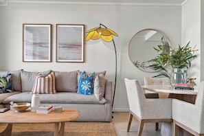 Ein Wohnzimmer mit Sofa, Couchtisch und Essbereich mit Stuhl und Tisch. An der Wand hängen zwei gerahmte Bilder, eine Stehlampe und ein Spiegel.