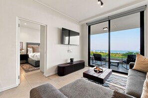 A modern living room with a large flat-screen TV, a sofa, a coffee table, and a view of the ocean through floor-to-ceiling glass doors. - Stylish Umhlanga 2Bed | Aircon and Gym (Umhlanga)
