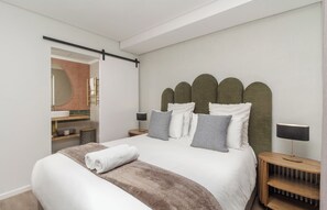 A modern bedroom with a large bed, a headboard with a unique design, bedside lamps, and a wooden nightstand. - Elegant De Waterkant Stay | Pool and Gym (Cape Town)