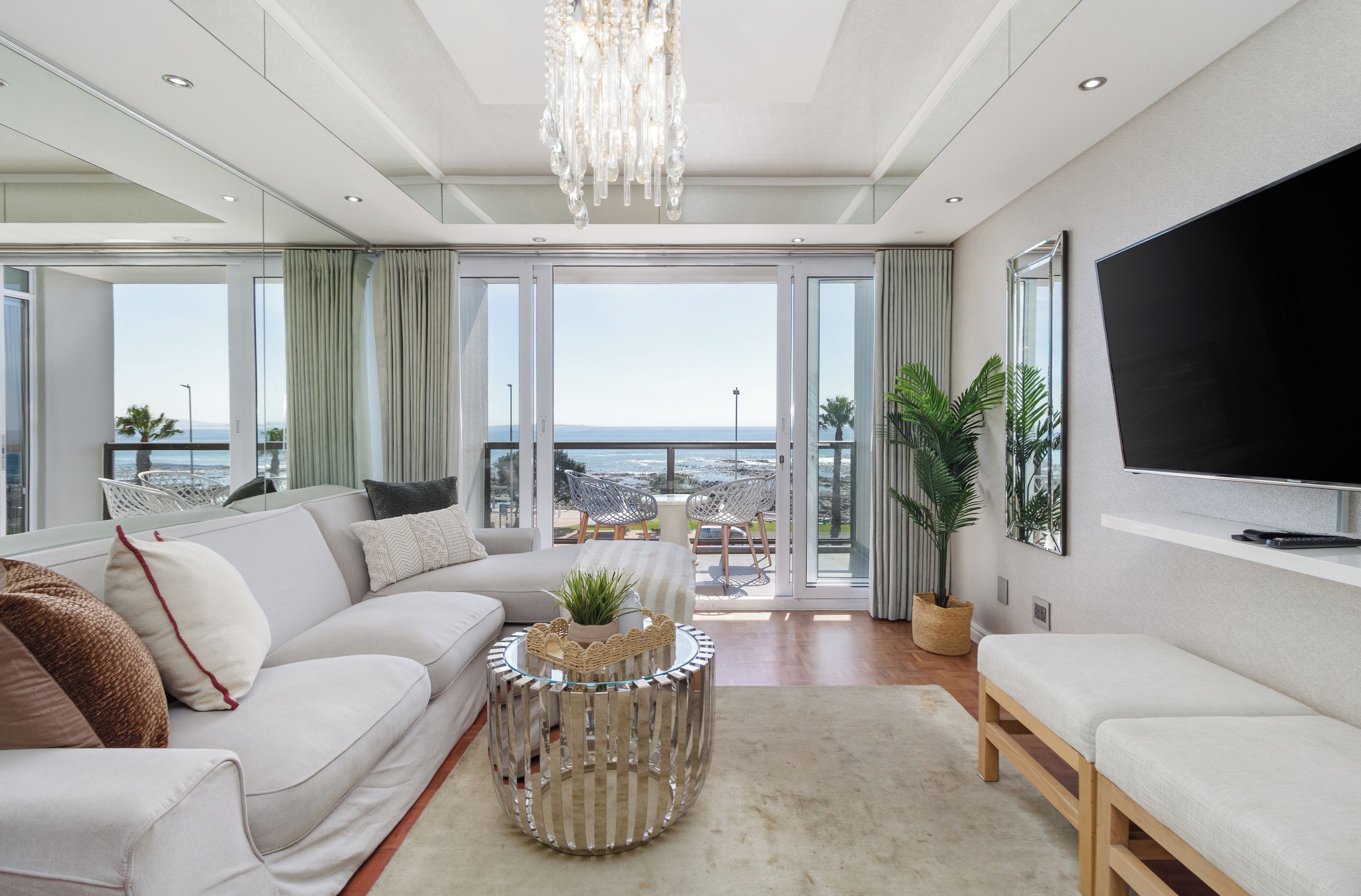 A modern living room with a sofa, a coffee table, and a TV. There is a balcony with a view of the sea.