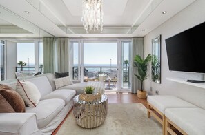 A modern living room with a sofa, a coffee table, and a TV. There is a balcony with a view of the sea. - Stylish Seaside Stay | Ocean Views and Balcony (Cape Town)