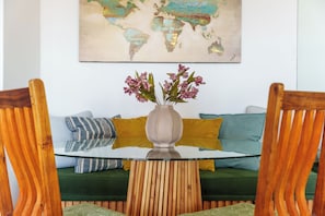 A round glass table with a vase of flowers, flanked by wooden chairs, in front of a green sofa with patterned cushions. - Stylish Seaside Stay | Ocean Views and Balcony (Cape Town)