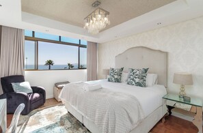 A bedroom with a large bed, a chair, a nightstand, and a window with a view. - Stylish Seaside Stay | Ocean Views and Balcony (Cape Town)
