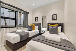 Ein Hotelzimmer mit zwei Betten, einem großen Fenster mit Jalousien, einem Spiegel und gerahmten Kunstwerken an den Wänden.