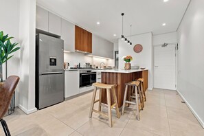 Une cuisine moderne avec un réfrigérateur en acier inoxydable, des armoires en bois, un îlot central doté de tabourets et une horloge au mur.